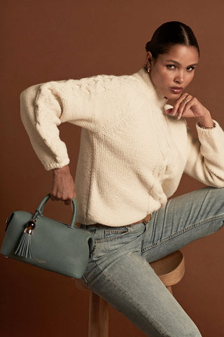 Female model holding Alessia Steel Green faux leather Bowler Tote by Luella Grey by the grab handles at the top of the bag