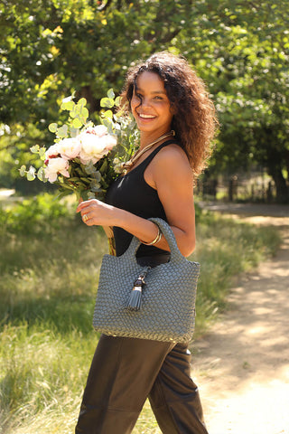 Female model wearing Suky Steel Green faux leather Woven Crossbody over her arm using the grab handles and holding flowers in the other hand