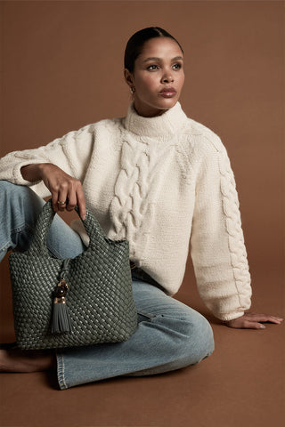Female model sat down holding Suky Steel Green faux leather Woven Crossbody by the top grab handles