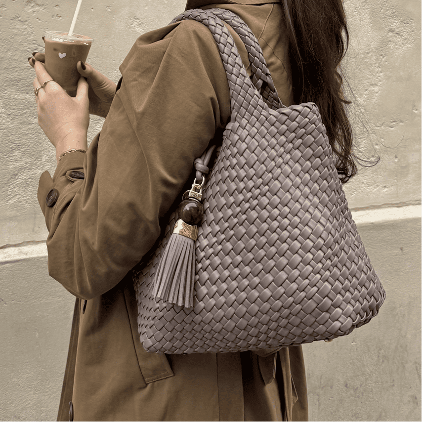Person holding a woven handbag with a tassel, wearing a brown coat against a neutral background